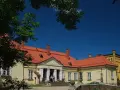 Pałac w Waplewie Wielkim, żółty, z czerwonym dachem, otoczony zielenią, pod błękitnym niebem.