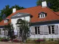 Jasnoniebieski dworek Sierakowskich z czerwoną dachówką, otoczony zielenią i ogrodzeniem.