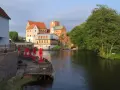 Zamek Darłowo z wieżami i zielonymi dachami, otoczony wodą i drzewami. Przy brzegu letnia restauracja z czerwonymi parasolami.