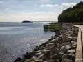 Kamieniste wybrzeże w Gdyni Oksywie z zielonym pomostem i falochronem. W oddali widać most i budynek na wodzie.