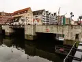 Kamienny most nad ciemną wodą, z rzędem zabytkowych kamienic w tle. W oddali widać dźwigi budowlane.