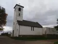 Biały, zabytkowy dwór z wieżą i dachem krytym dachówką. W oddali widać bramę i drzewa.