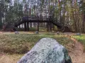 Kamienne kręgi w Odrach. Drewniana kładka nad leśnym wzgórzem, otoczona głazami i sosnami. Na pierwszym planie kamień z monetami.