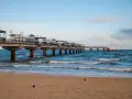 Molo w Międzyzdrojach, jedna z głównych atrakcji, wznosi się nad wzburzonym morzem. Piasek plaży i błękitne niebo tworzą malowniczy krajobraz.