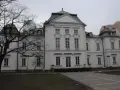 Pałac Przebendowskich, biała, elegancka budowla z zielonym dachem, otoczona drzewami i nowoczesnymi budynkami.