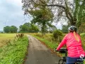 Kobieta na rowerze jedzie starym kolejowym szlakiem wśród zieleni i drzew. W oddali widać wiatraki.