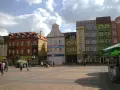 Rynek w Chojnicach z kolorowymi kamienicami, w tym ratusz. Ludzie spacerują po brukowanym placu.