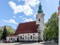 Biała bazylika pw. NMP Królowej Polski z czerwoną dachówką i zieloną wieżą, otoczona drzewami i budynkami.