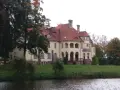 Malowniczy pałac z czerwoną, dachówkową, spadzistą dachem, otoczony zielenią i wodą.