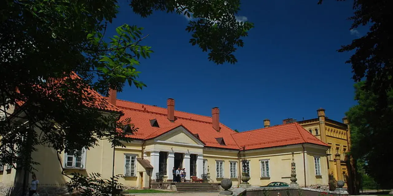 Pałac w Waplewie Wielkim, żółty, z czerwonym dachem, otoczony zielenią, pod błękitnym niebem.