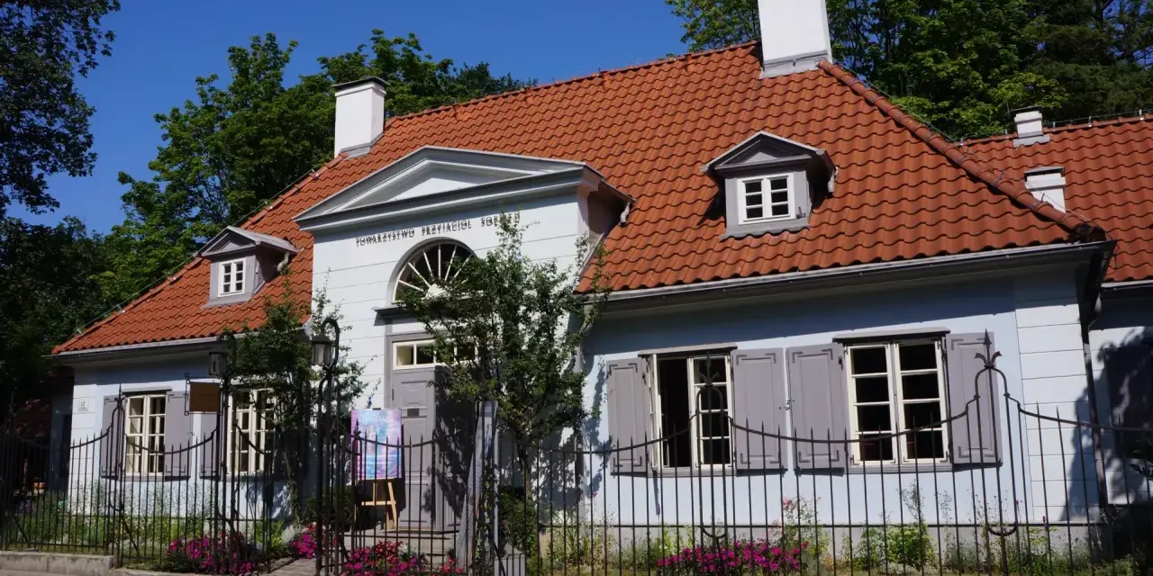 Jasnoniebieski dworek Sierakowskich z czerwoną dachówką, otoczony zielenią i ogrodzeniem.