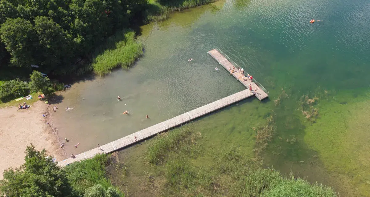 Letnie popołudnie nad jeziorem Hańcza. Ludzie kąpią się i odpoczywają na plaży, inni spacerują po drewnianym pomoście.