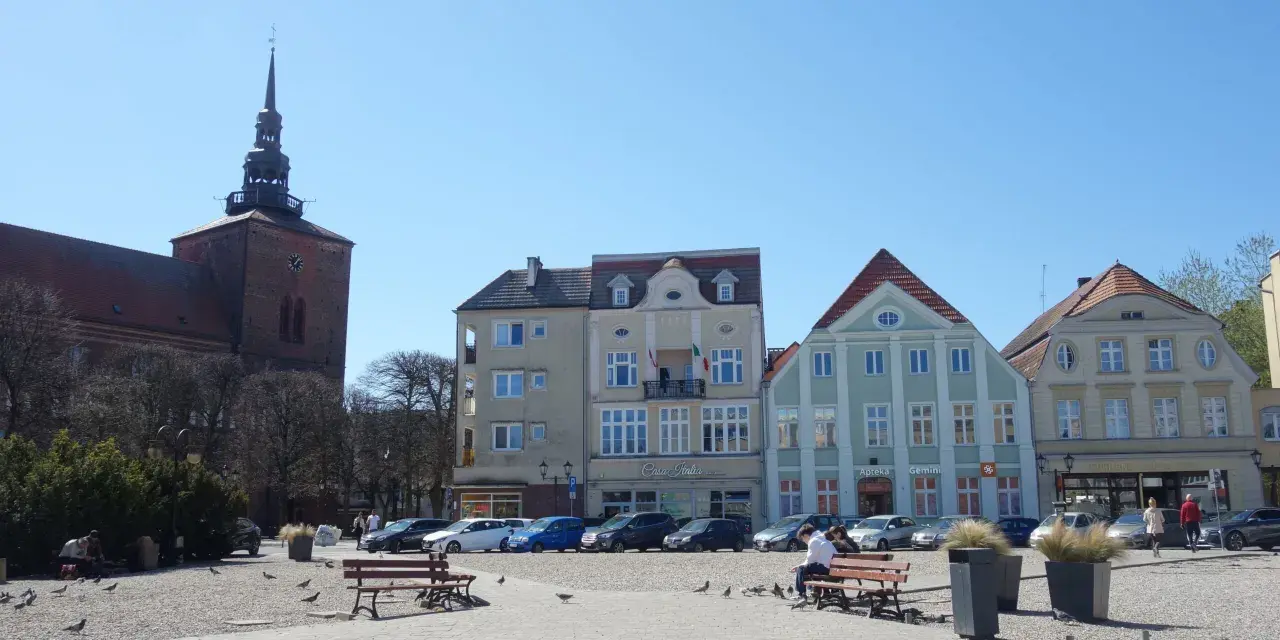Słupsk, stary rynek: kościół z wieżą, kolorowe kamienice, ławki, ludzie i gołębie na brukowanym placu.