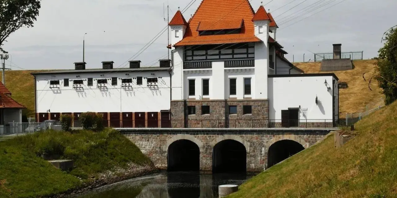 Elektrownia wodna Straszyn z charakterystycznym czerwonym dachem i kamiennymi arkadami nad kanałem.