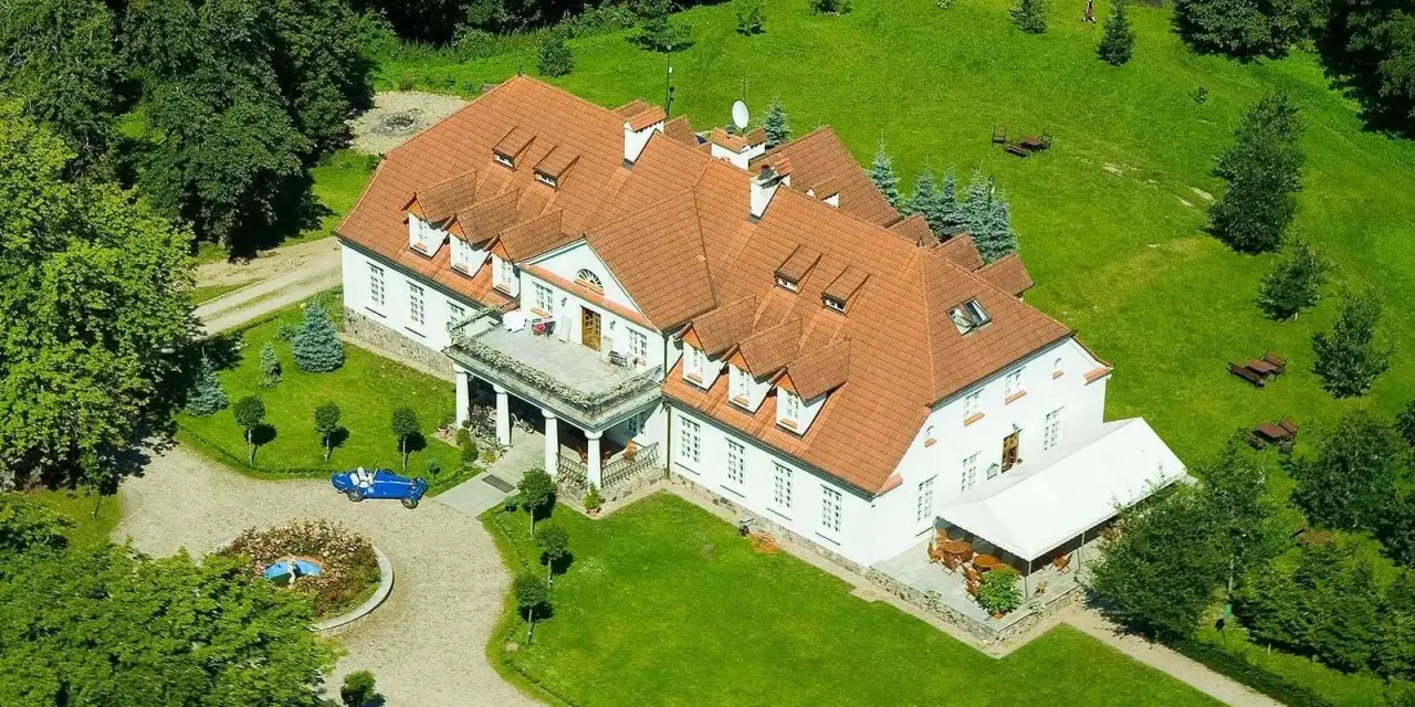 Dwór w Bychowie otoczony zielenią, z niebieskim kabrioletem przy różanym klombie.