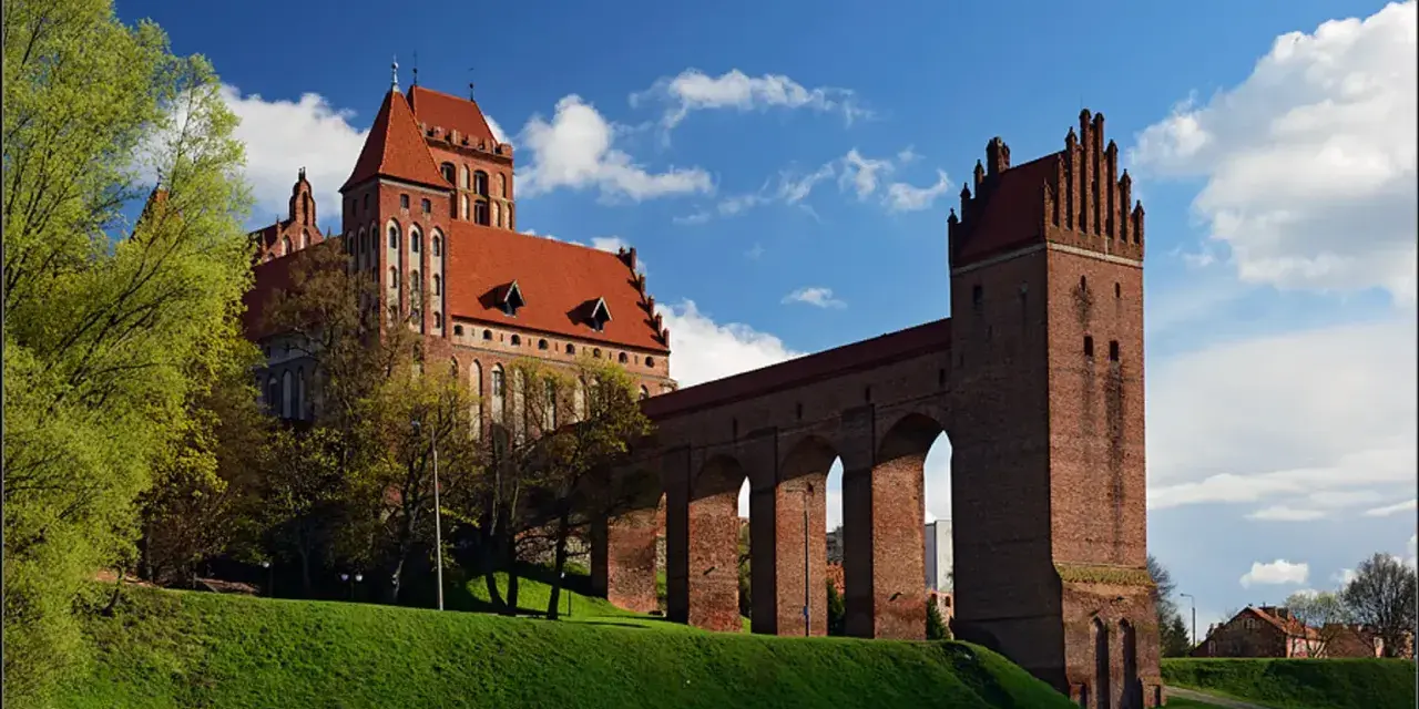 Majestatyczny zamek w Kwidzynie, z czerwonej cegły, z charakterystycznym gdaniskiem i długim, arkadowym murem obronnym, wznosi się na zielonym wzgórzu pod błękitnym niebem.