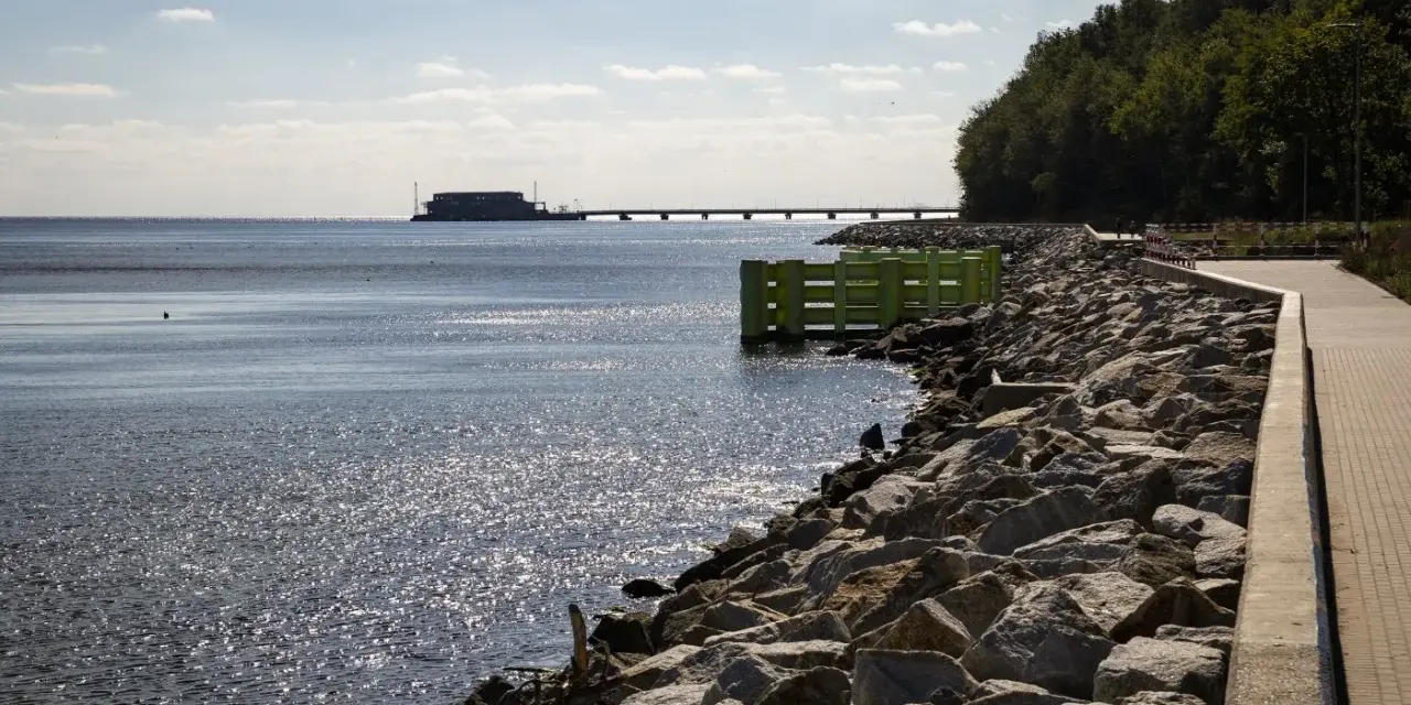 Kamieniste wybrzeże w Gdyni Oksywie z zielonym pomostem i falochronem. W oddali widać most i budynek na wodzie.