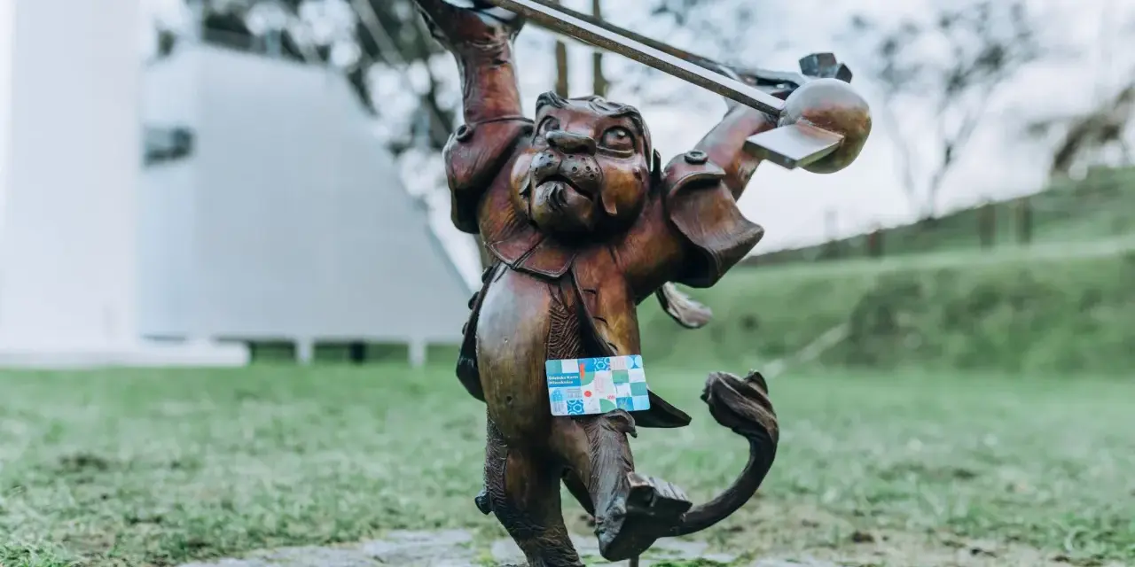 Figurka lew hewelion z kartą miejską w łapie, stojąca na bruku wśród zieleni.