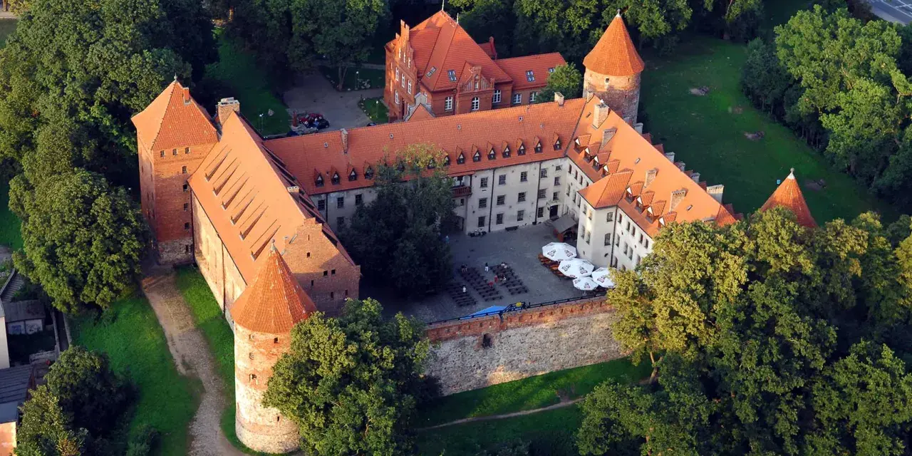 Zamek w Bytowie otoczony zielenią, z czerwonymi dachami i okrągłymi wieżami. Na dziedzińcu widać stoliki i parasole.