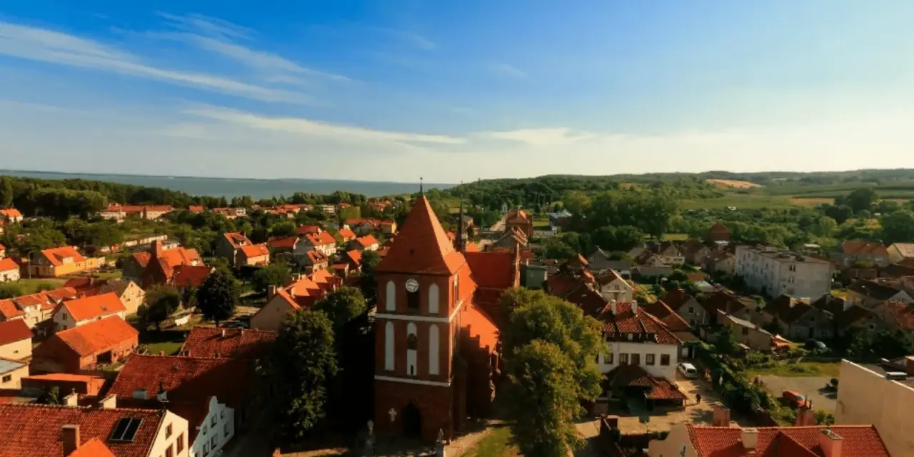 Widok na urokliwe Tolkmicko z charakterystyczną wieżą kościelną. Wśród czerwonych dachów i zieleni rozciąga się malowniczy krajobraz, idealny na odkrywanie tolkmickich atrakcji.
