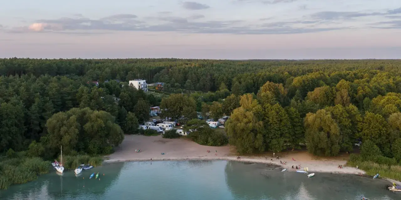 Plaża nad najczystszym jeziorem w Polsce, otoczona lasem. Woda jest turkusowa, na piasku widać ludzi i kampery.