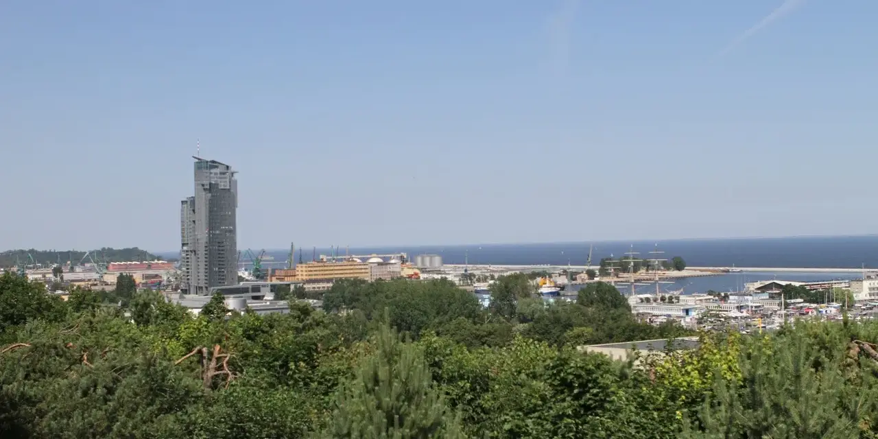 Widok na port w Gdyni z dominującym wieżowcem. W oddali morze, a na pierwszym planie zieleń drzew. Kamienna Góra w Gdyni oferuje takie panoramy.