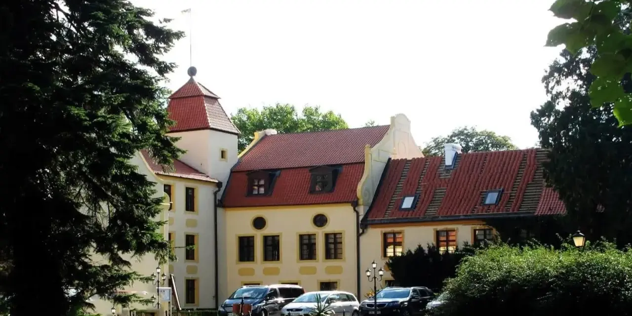 Zamek w Krokowej z czerwoną wieżą i dachami, otoczony zielenią. Przed budynkiem zaparkowane samochody.