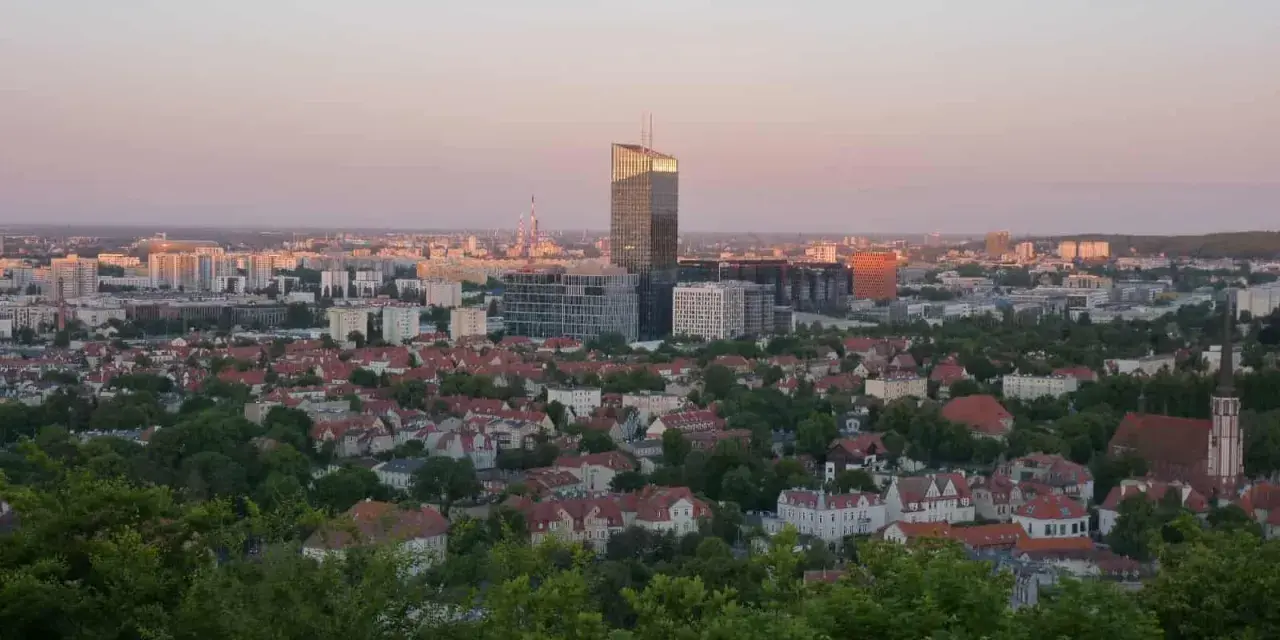 Widok na miasto o zachodzie słońca. W oddali widać nowoczesne budynki, a bliżej dachy domów i zieleń. Jakby pachołek oliwa na tle nieba.