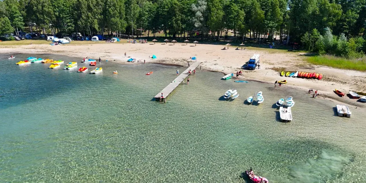 Letnia przygoda na wodzie, kolorowe rowerki wodne i kajaki czekają przy brzegu. Widok na plażę z namiotami i lasem, idealne miejsce na wypoczynek nad jeziorem Binduga 69.