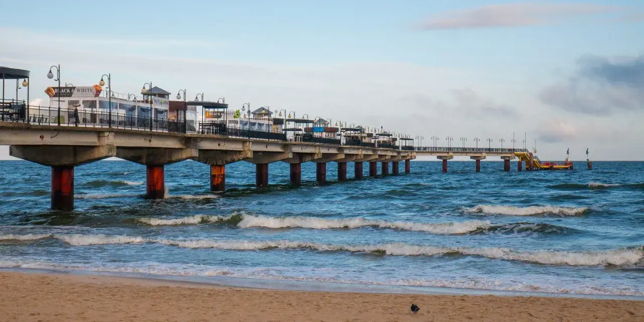 Molo w Międzyzdrojach, jedna z głównych atrakcji, wznosi się nad wzburzonym morzem. Piasek plaży i błękitne niebo tworzą malowniczy krajobraz.