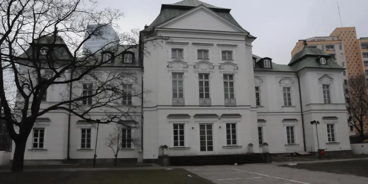 Pałac Przebendowskich, biała, elegancka budowla z zielonym dachem, otoczona drzewami i nowoczesnymi budynkami.