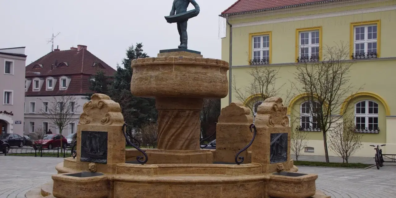 Kamienna fontanna z rzeźbą rybaka na szczycie, ozdobiona płaskorzeźbami statków.