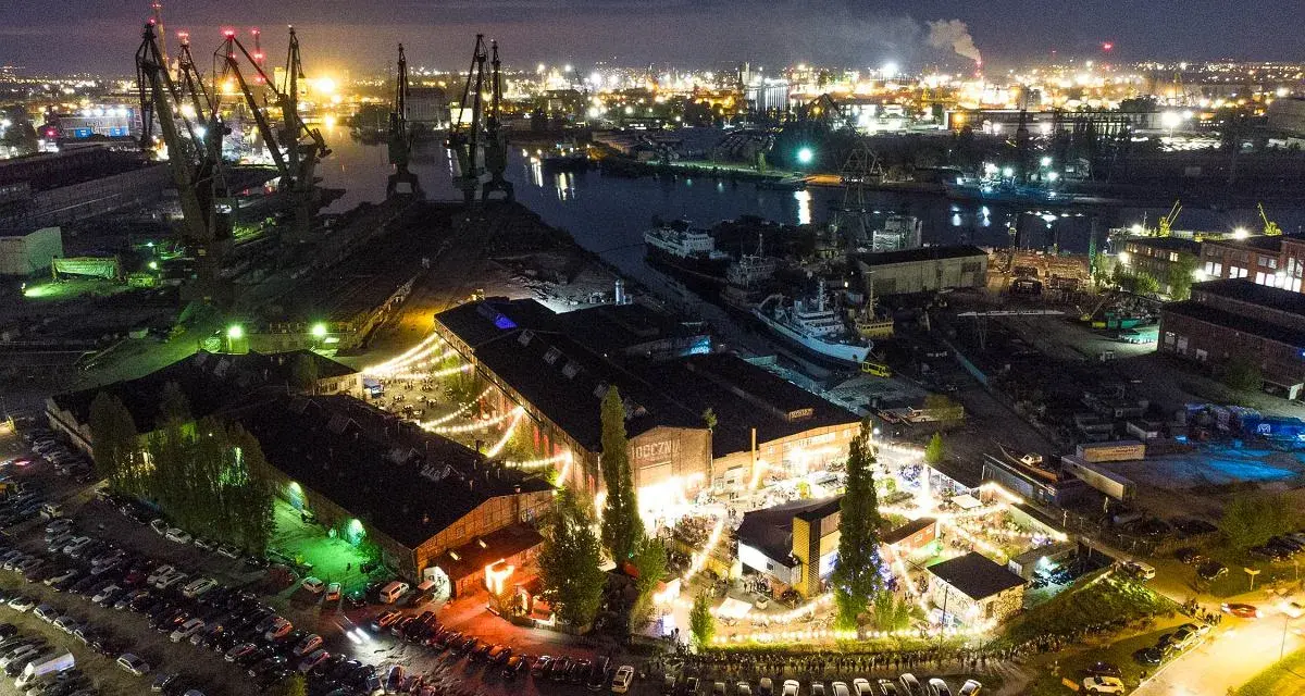 Nocny widok na stocznię w Gdańsku, gdzie odbywa się wydarzenie. Widać dźwigi, statki i budynki oświetlone lampkami. W pobliżu parkują samochody, jakby przyjechali tu elektrycy z Gdańska na imprezę.