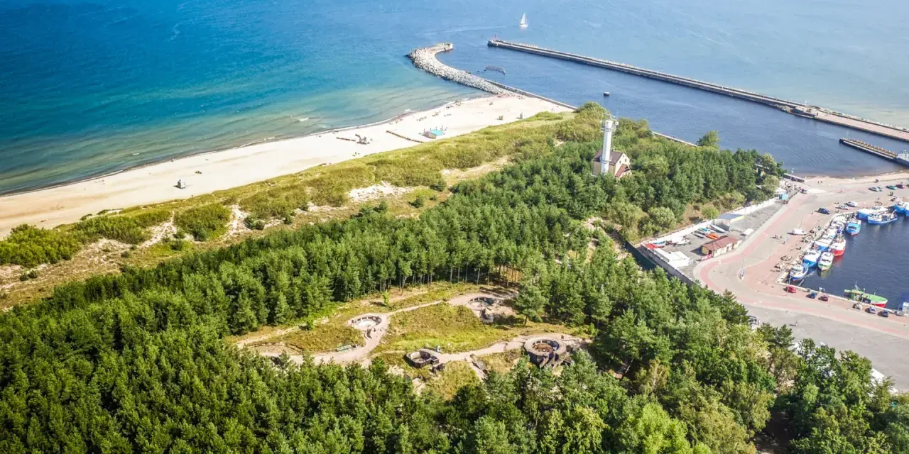 Widok z lotu ptaka na wybrzeże z piaszczystą plażą, lasem i portem. Wśród drzew widoczne są historyczne bunkry nad morzem.