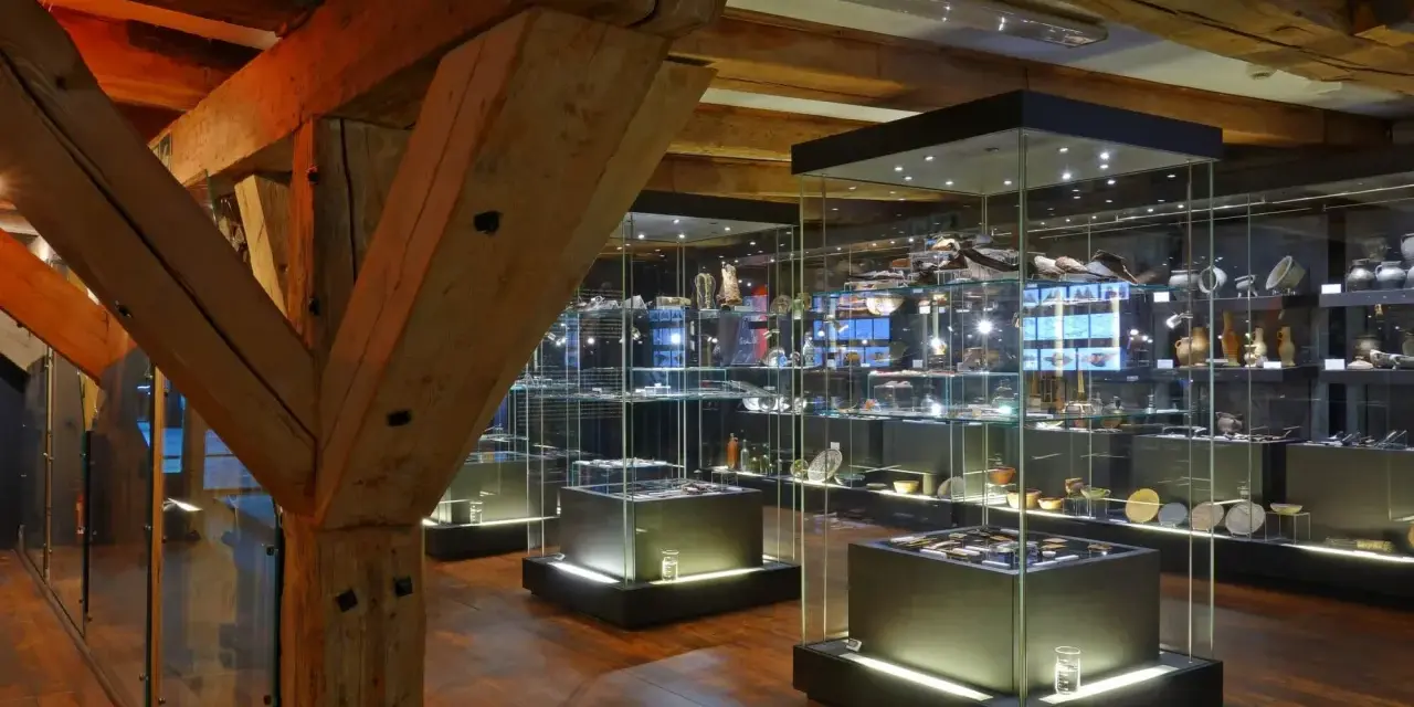 Wnętrze muzeum z zabytkowymi belkami i gablotami pełnymi artefaktów. Wśród nich można odnaleźć spichlerz błękitny baranek, symbol dawnych czasów.