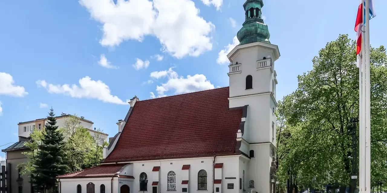 Biała bazylika pw. NMP Królowej Polski z czerwoną dachówką i zieloną wieżą, otoczona drzewami i budynkami.