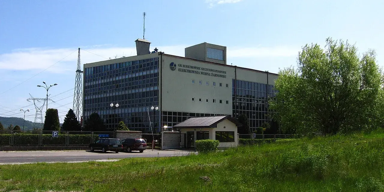 Budynek elektrowni wodnej Żarnowiec z charakterystycznym logo. W tle wieża transmisyjna i zieleń.