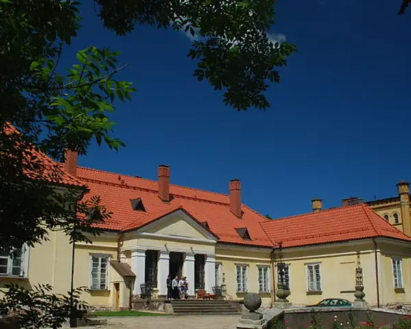 Pałac w Waplewie Wielkim, żółty, z czerwonym dachem, otoczony zielenią, pod błękitnym niebem.