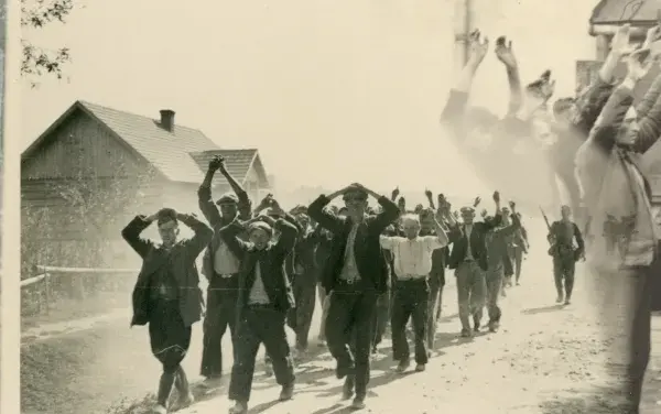 Zatrzymani mężczyźni z rękami w górze na drodze, być może w rejonie granica polsko-niemiecka 1939 Pomorze. W tle domy i żołnierz.