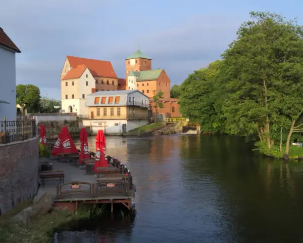 Zamek Darłowo z wieżami i zielonymi dachami, otoczony wodą i drzewami. Przy brzegu letnia restauracja z czerwonymi parasolami.