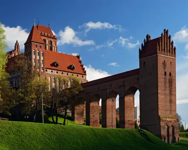 Majestatyczny zamek w Kwidzynie, z czerwonej cegły, z charakterystycznym gdaniskiem i długim, arkadowym murem obronnym, wznosi się na zielonym wzgórzu pod błękitnym niebem.
