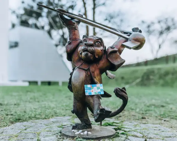 Figurka lew hewelion z kartą miejską w łapie, stojąca na bruku wśród zieleni.