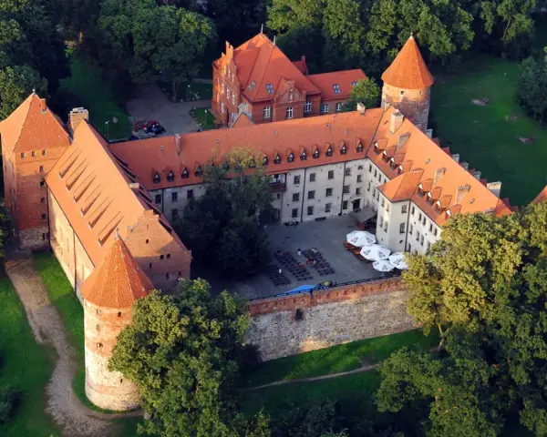 Zamek w Bytowie otoczony zielenią, z czerwonymi dachami i okrągłymi wieżami. Na dziedzińcu widać stoliki i parasole.