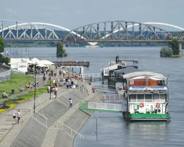 Ludzie spacerują bulwarem nad Wisłą, podziwiając widok na mosty. W oddali widać statek-restaurację, a na brzegu kawiarnię z lodami.
