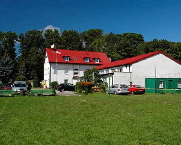 Budynek z czerwonym dachem, przypominający dawny korsarz darłówko, otoczony zielenią i samochodami.