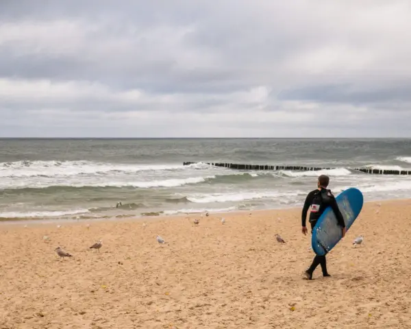 Surfer idzie plażą w Juracie, szukając fal. To jedna z wielu atrakcji tego miejsca.