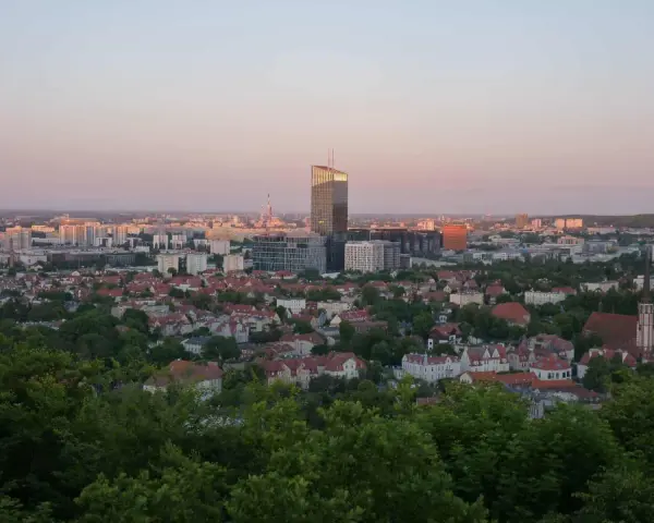 Widok na miasto o zachodzie słońca. W oddali widać nowoczesne budynki, a bliżej dachy domów i zieleń. Jakby pachołek oliwa na tle nieba.
