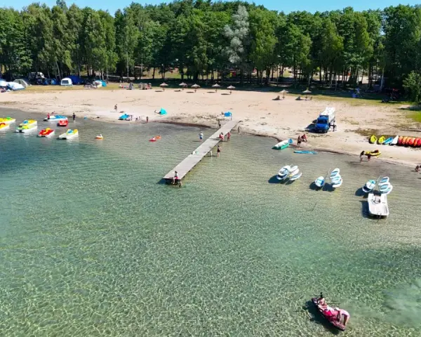 Letnia przygoda na wodzie, kolorowe rowerki wodne i kajaki czekają przy brzegu. Widok na plażę z namiotami i lasem, idealne miejsce na wypoczynek nad jeziorem Binduga 69.