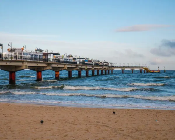 Molo w Międzyzdrojach, jedna z głównych atrakcji, wznosi się nad wzburzonym morzem. Piasek plaży i błękitne niebo tworzą malowniczy krajobraz.