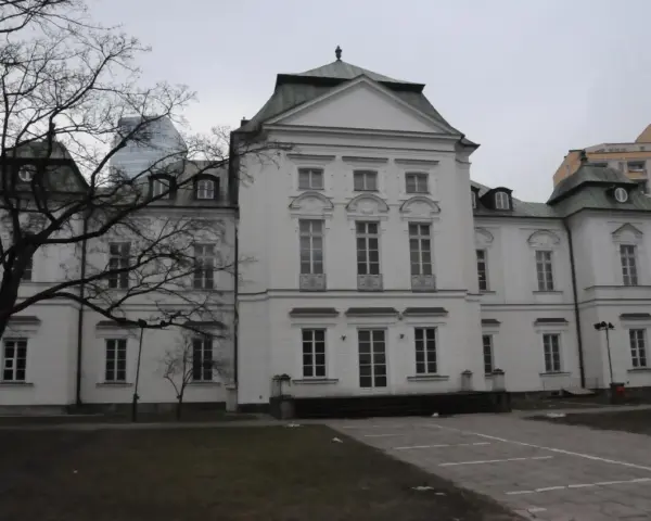 Pałac Przebendowskich, biała, elegancka budowla z zielonym dachem, otoczona drzewami i nowoczesnymi budynkami.